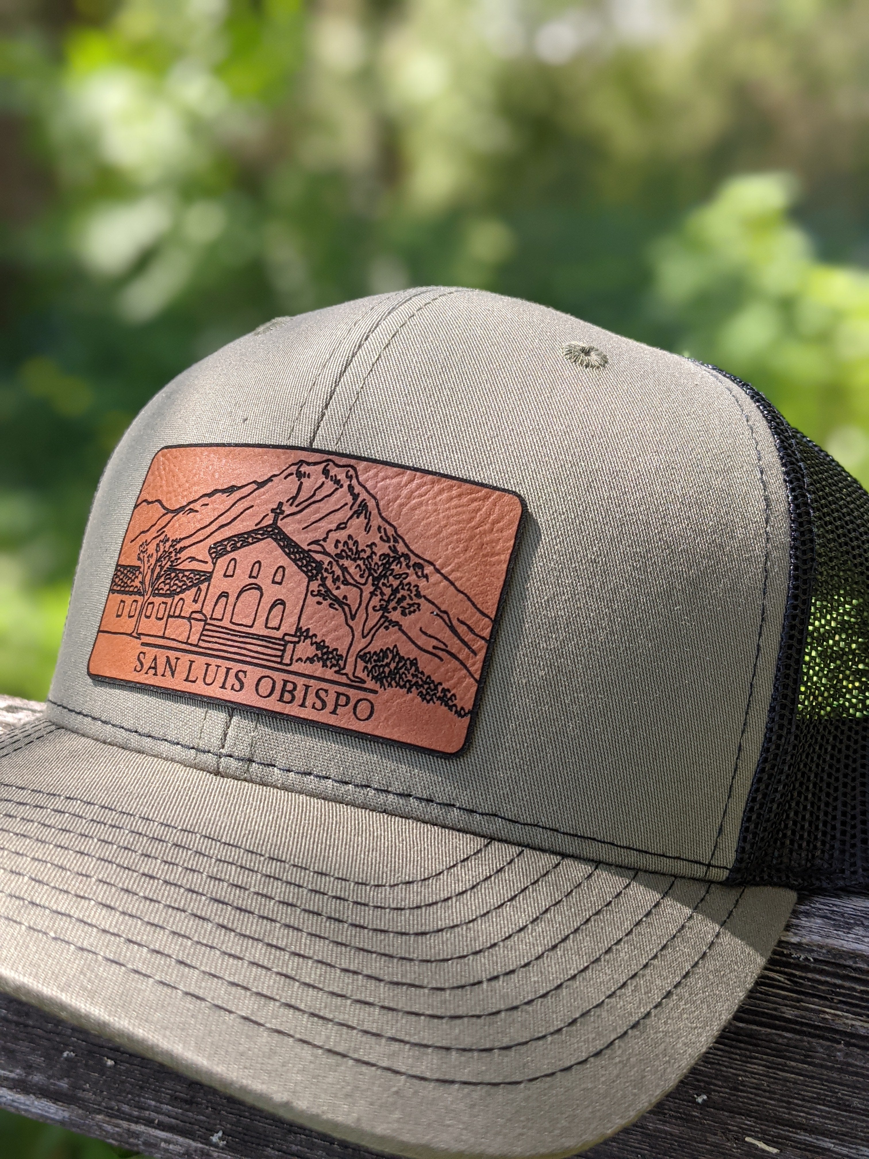 Trucker hat with black mesh on back, olive green colored front and leather patch depicting the Mission and Madonna Mountain. The patch reads "San Luis Obispo."